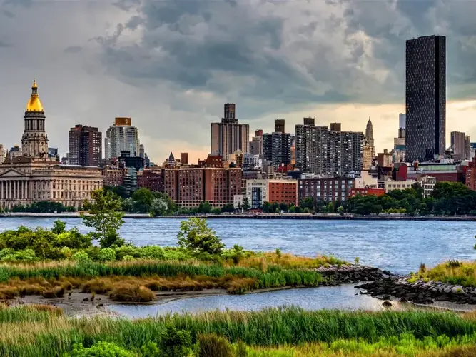 A city skyline with a body of water in the foreground and a cloudy sky above.